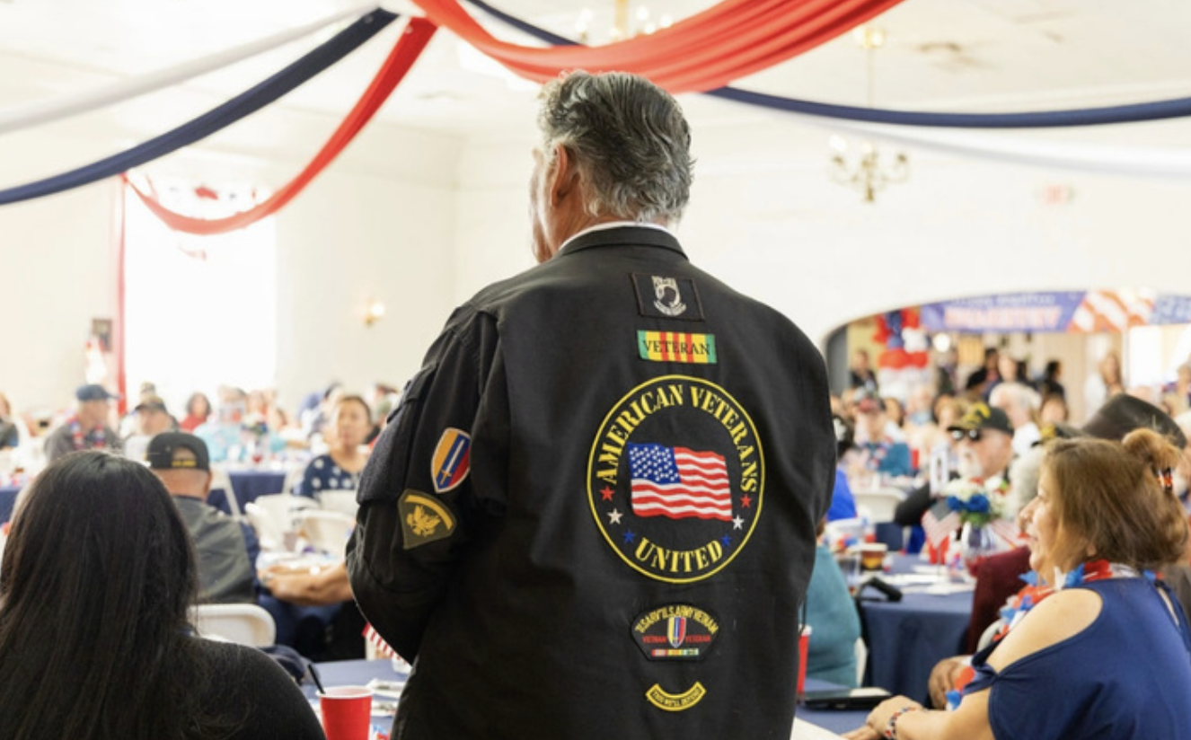 A man wearing a black jacket with patches, including a large circular one that reads th American Veterans United with an American flag, is standing at a patriotic event with red, white, and blue decorations. The event appears to be attended primari