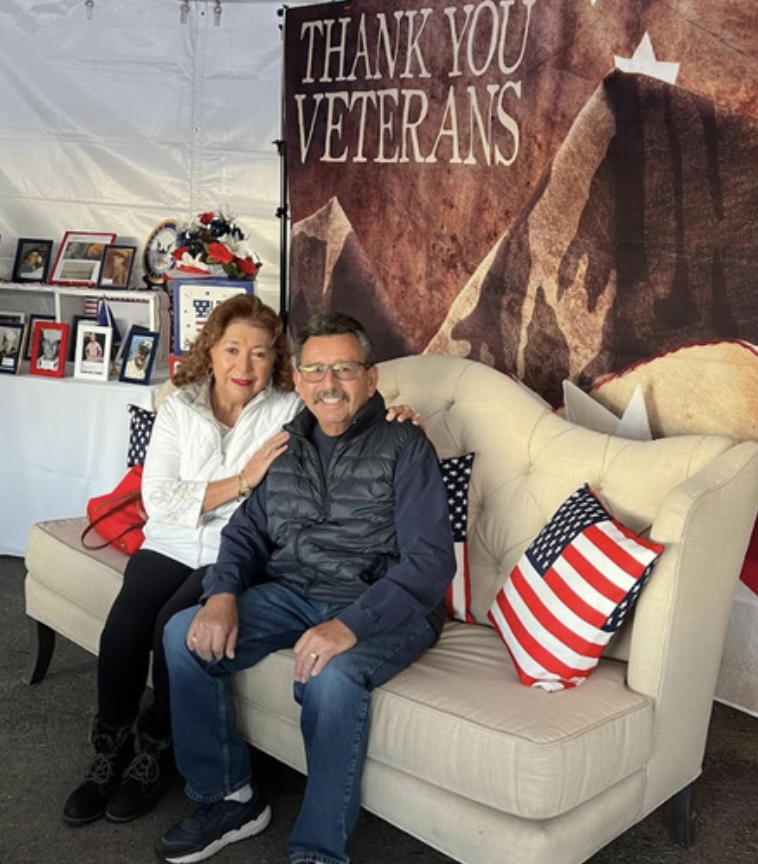 A man and woman sitting on a beige couch under a large sign that says 'Thank You Veterans'. The woman has red curly hair, and the man is wearing glasses and a black vest. There are American flag pillows on the couch and a display of framed photos and