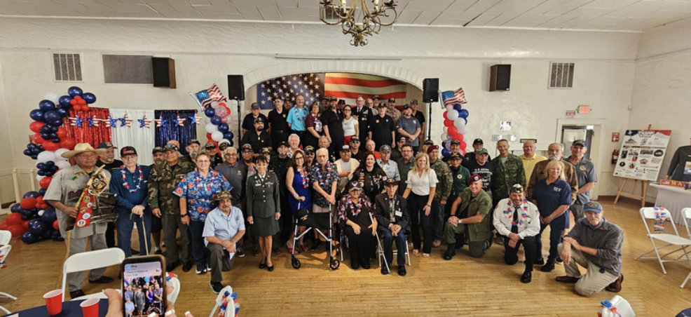 Large group of people, including military personnel and civilians, gathered in a decorated room with American flags and red, white, and blue balloons, celebrating a patriotic event.