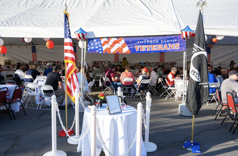 A tent decorated for a veterans appreciation event with tables, people sitting, American flags, and a banner that says "Thank you VETERANS."