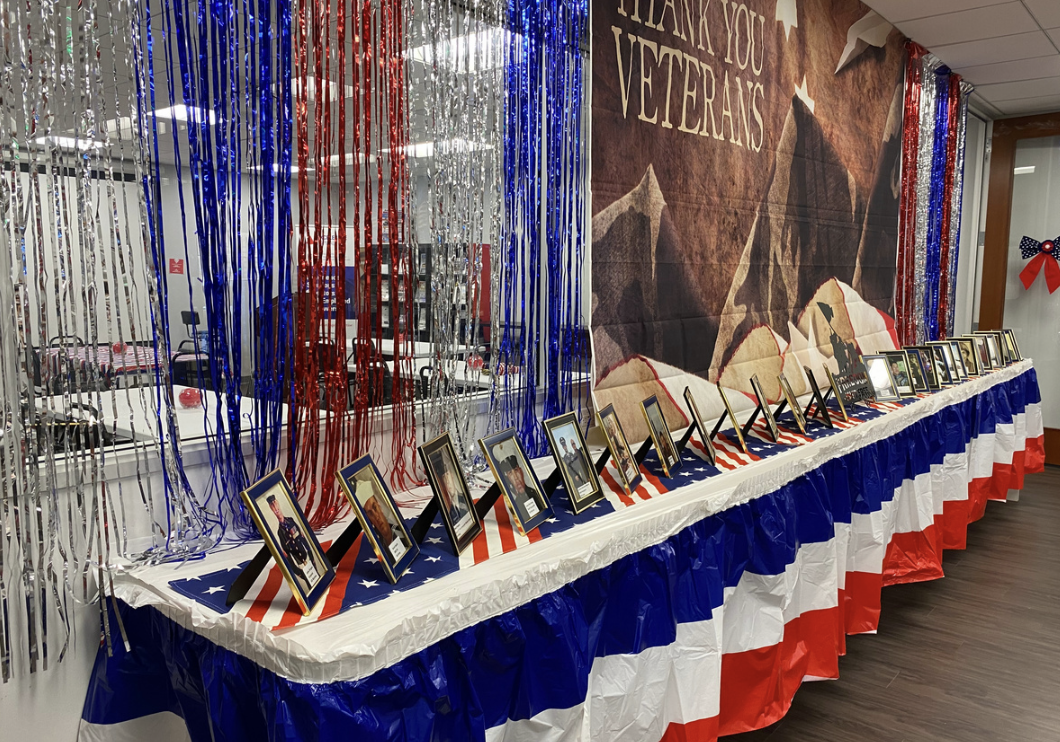 Table decorated with red, white, and blue patriotic colors, displaying framed photographs of veterans, with a large banner behind that says 'Thank You Veterans' and tinsel streamers in red, white, blue, and silver.