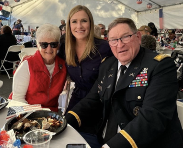 Three people sitting at a table under a tent at an outdoor celebration, with an elderly woman in a red vest, a young woman, and a man in a military uniform with medals. They are smiling, and there are plates of food and decorations on the table.