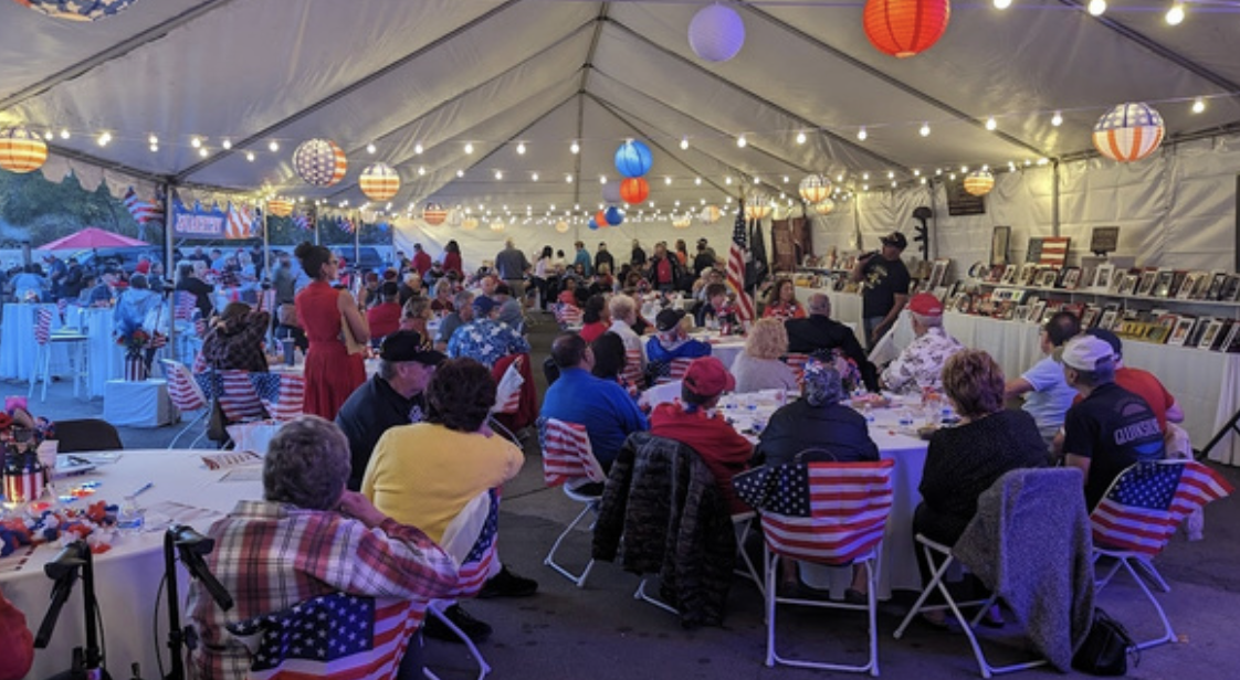 Patriotic-themed gathering inside a large tent with American flags, red, white, and blue decorations, and a crowd of people sitting at decorated tables.