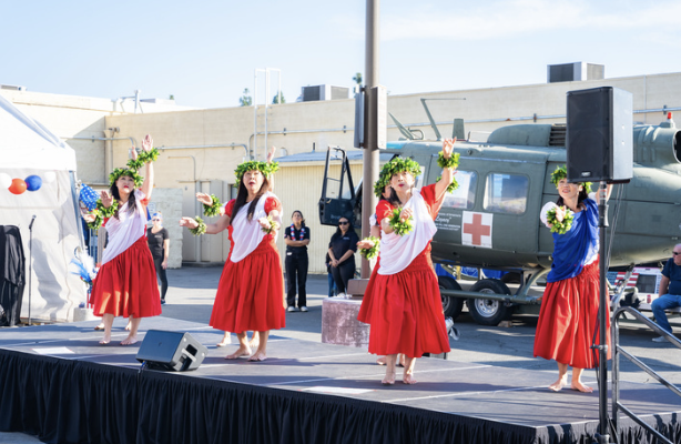 Four women in red skirts and white tops performing a traditional dance on an outdoor stage, wearing floral headpieces, with a helicopter and people watching in the background.