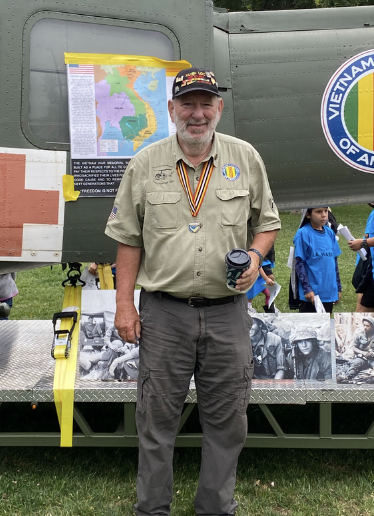 A man in a tan military or veteran shirt with medals and a cap, holding a cup, standing outdoors in front of a helicopter and historical photos during an event. Other people and a Vietnam map are visible in the background.
