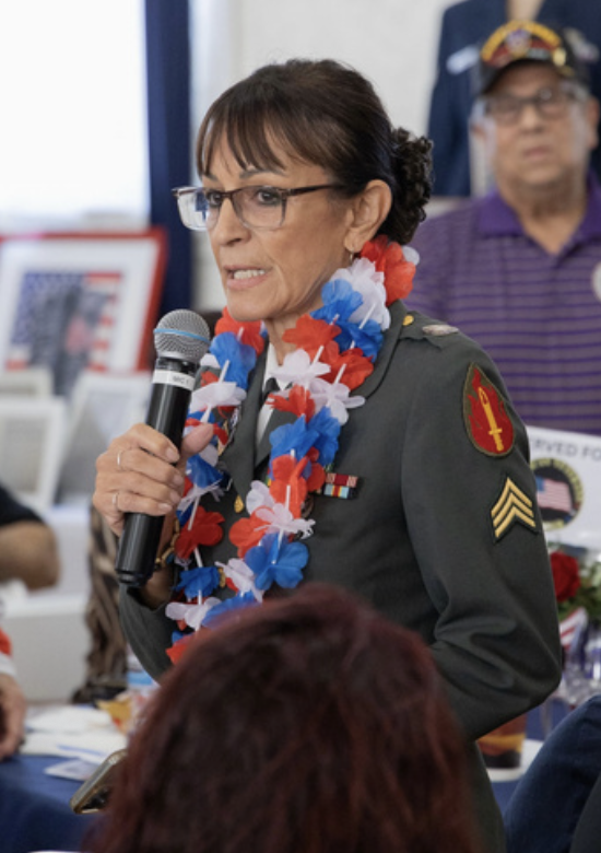 A woman in military uniform speaking into a microphone wearing a red, white, and blue lei. A man in a purple striped shirt and glasses stands behind her.