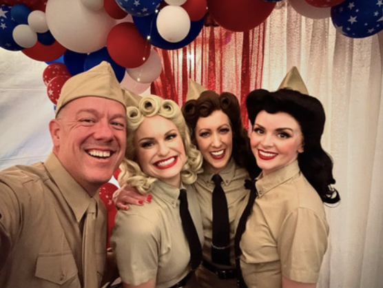 Four people dressed as characters from the movie 'The Wizard of Oz' in front of red, white, and blue balloons and patriotic decorations.