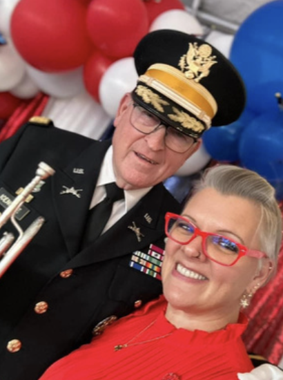A man dressed in a military uniform with medals and a hat, and a woman wearing red glasses and a red dress, taking a close-up selfie together with balloons in the background.
