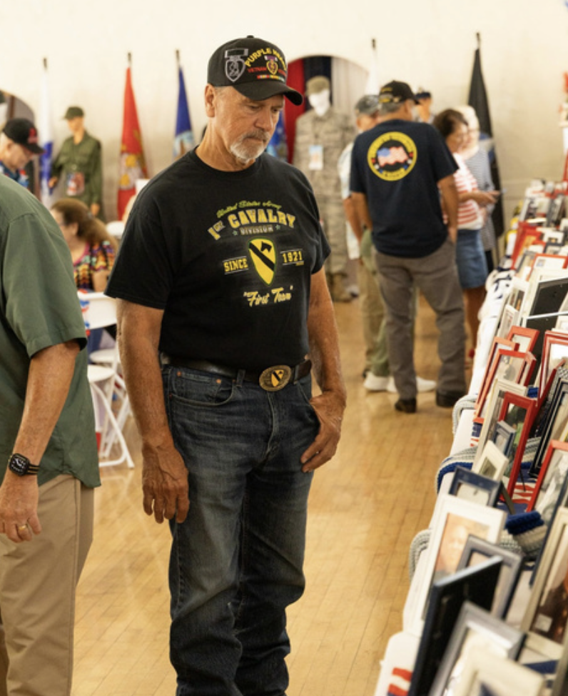 A man in a Black Veterans of Foreign Wars cap and a T-shirt with a military-related logo looks at a display of framed photographs at an indoor event surrounded by other attendees.