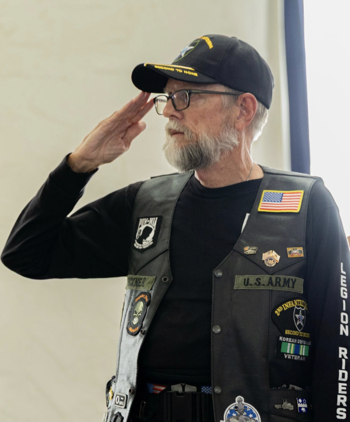 An elderly man with a beard and glasses salutes wearing a U.S. Army vest and a baseball cap, with various military patches and insignia.
