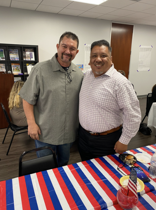 Two men standing together, smiling, at a table with red, white, and blue striped tablecloth in a room with bookshelves and other people seated.
