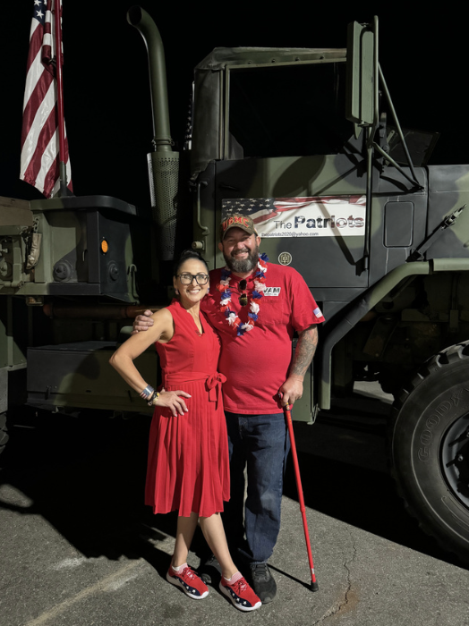 Two people standing in front of a military vehicle at night, both wearing red clothing and holiday accessories, smiling for the camera.