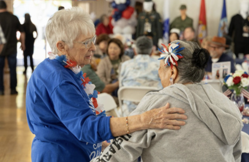 Two elderly women, one in a blue shirt and the other in a gray hoodie, are engaging in a friendly interaction at a patriotic-themed celebration, with several people sitting at tables in the background.