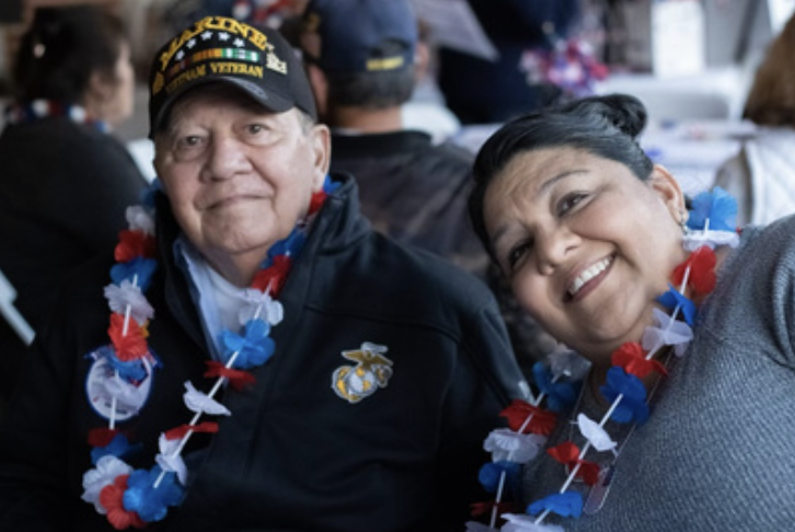 A man and woman at a patriotic event wearing red, white, and blue floral leis, with the man in a navy jacket with a Marine emblem and wearing a cap that says "BATTLE R honor the Marine Corps." The woman is smiling and leaning towards the camera.