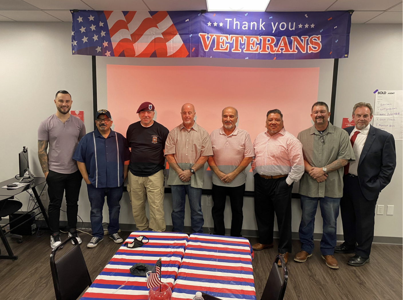 Group of nine men standing in a line in front of a backdrop with an American flag and a sign that says 'Thank you Veterans' at a celebration event. There is a table with a patriotic tablecloth and a small American flag in the foreground.