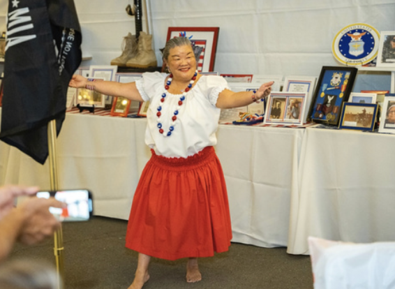 Woman dancing happily in a patriotic outfit with a white blouse and red skirt at an event with framed pictures and memorabilia on tables behind her.