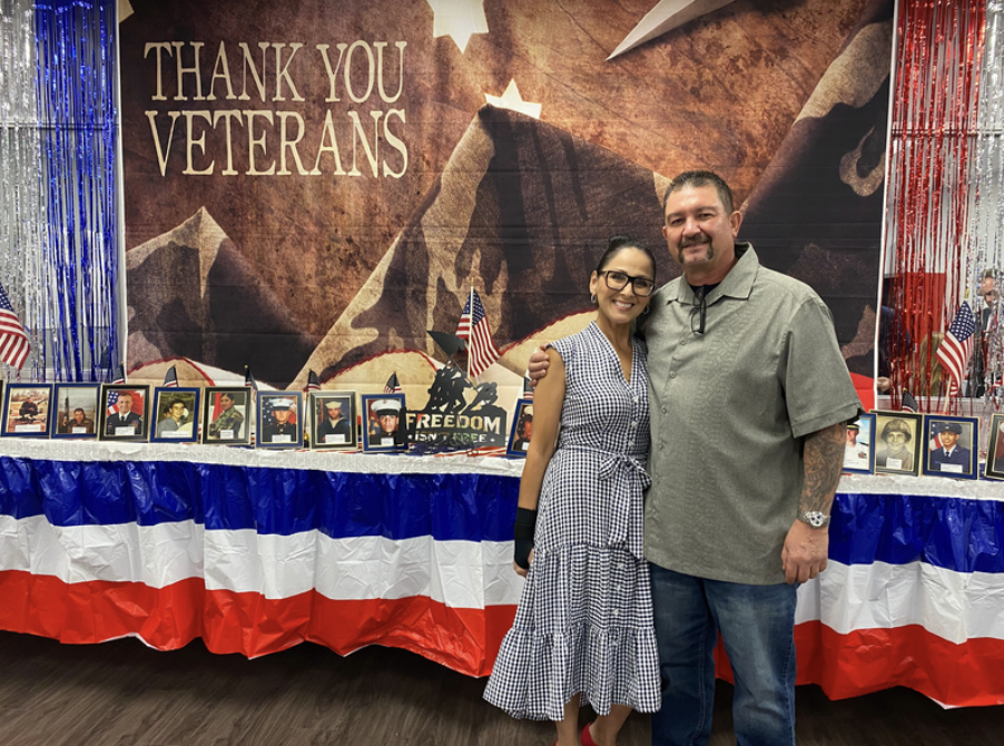 Two people standing together in front of a memorial display thanking veterans, with photos of veterans, American flags, and patriotic decorations.