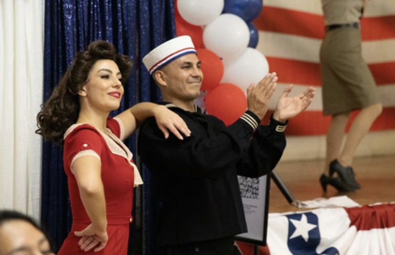 Two people dressed in vintage attire at a patriotic event, with the woman in a red dress and the man in a Navy hat clapping, and a large American flag decoration in the background.