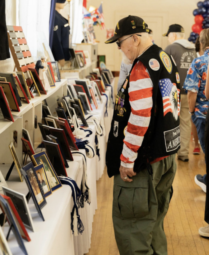 An elderly man wearing a military veteran vest and a cap is looking at framed photographs and medals displayed on a table at an indoor event honoring veterans.