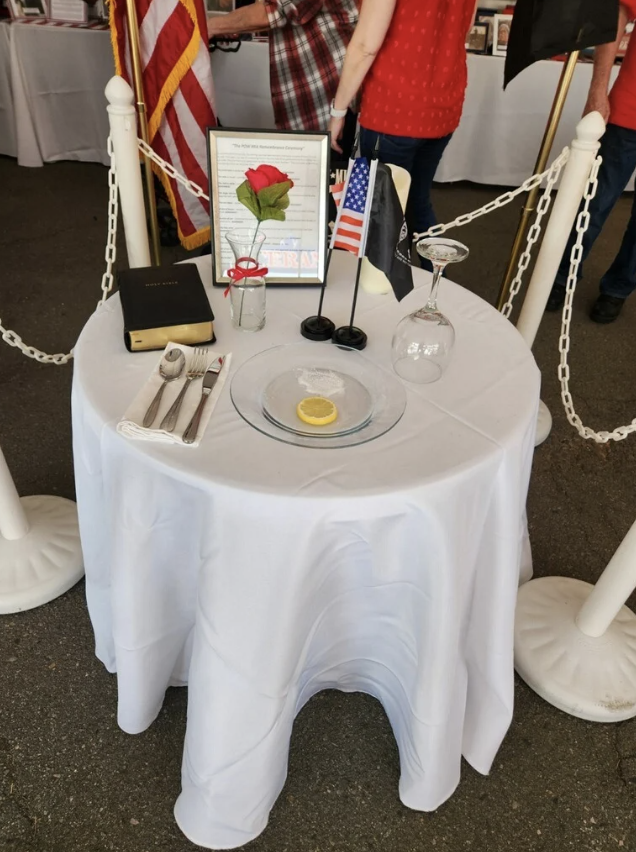 A ceremonial table set with a white tablecloth, a lemon slice on a clear glass plate, a flower in a small glass jar with a ribbon, a framed document, and flags of the United States and other entities. The table is surrounded by white posts with chains, located indoors with people and display tables in the background.