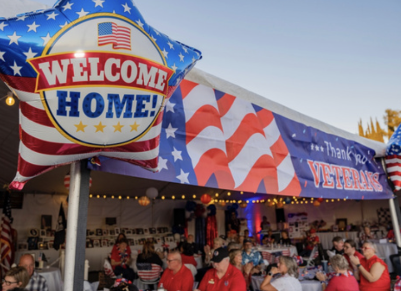 People gathered in a decorated tent celebrating Veterans Day with patriotic banners and a "Welcome Home!" balloon.