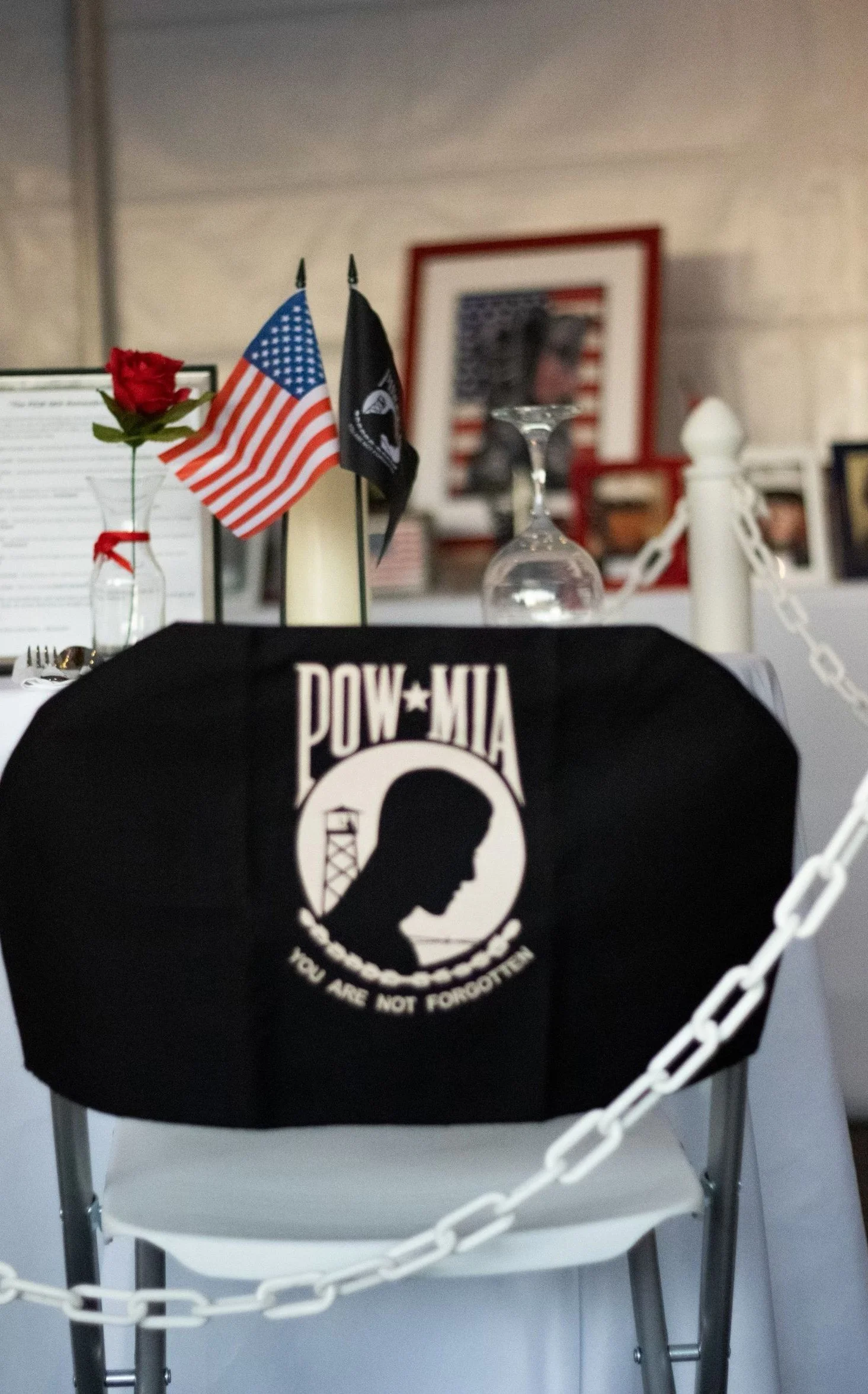 A black harness with the Pow Mia logo and the message "You Are Not Forgotten" hanging on a chair in front of a table with flags, a framed picture of the American flag, and a glass upside down.