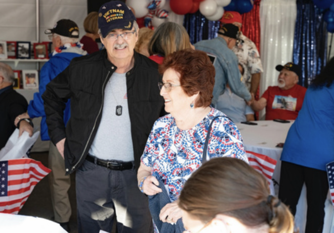 An elderly man and woman smiling and chatting at a social gathering with patriotic decorations, including red, white, and blue balloons and American flags in the background.