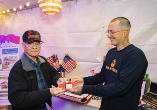 Two men exchanging a cake at an indoor event with American flags and patriotic decorations.