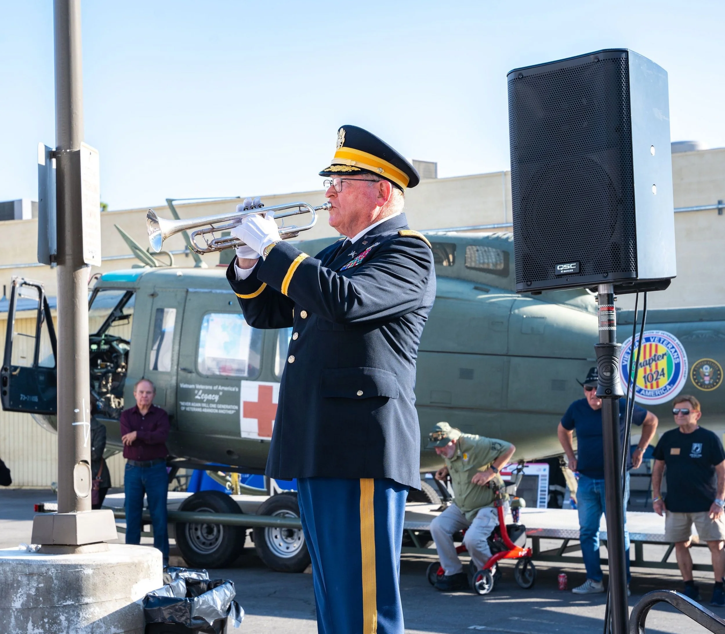 A man dressed in a military uniform playing a trumpet at an outdoor event, with a helicopter and several people in the background.