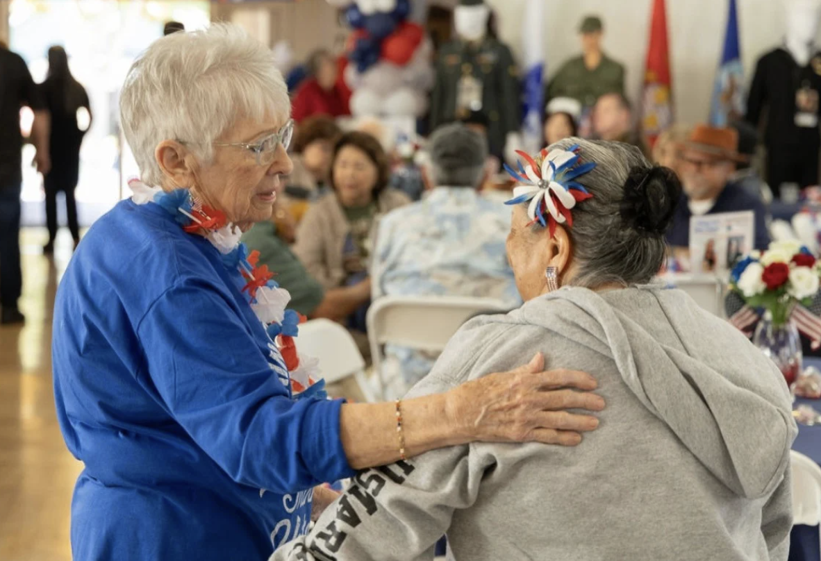 Two women wearing patriotic red, white, and blue decorations celebrate at a gathering, with one woman placing her hand on the other's shoulder.