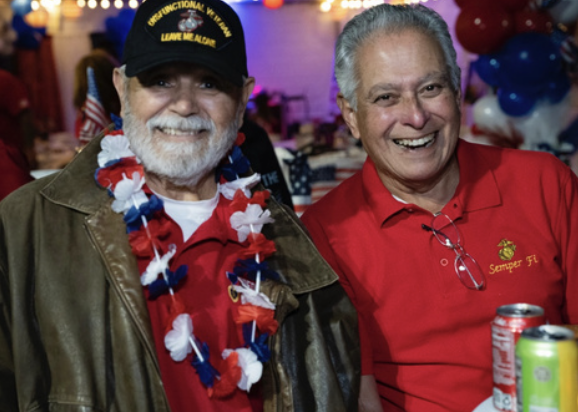 Two smiling men at a patriotic celebration, one wearing a black cap and a patriotic lei, the other in a red polo shirt, with balloons and decorations in the background.