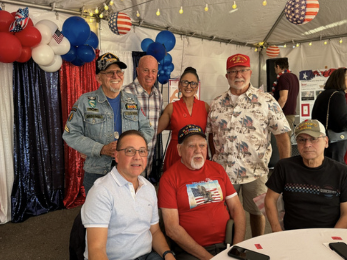 Group of six adults, four men and two women, sitting and standing inside a decorated tent with red, white, and blue balloons and American flag-themed decorations. They are at a patriotic celebration.