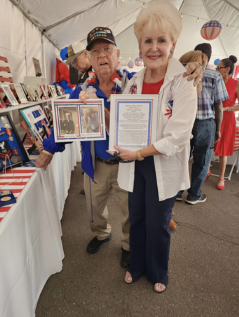 An elderly man and woman at an event, posing with framed photographs and a document. The man wears a magazine cap and patriotic attire, and the woman holds a framed document. There are people and display tables in the background.