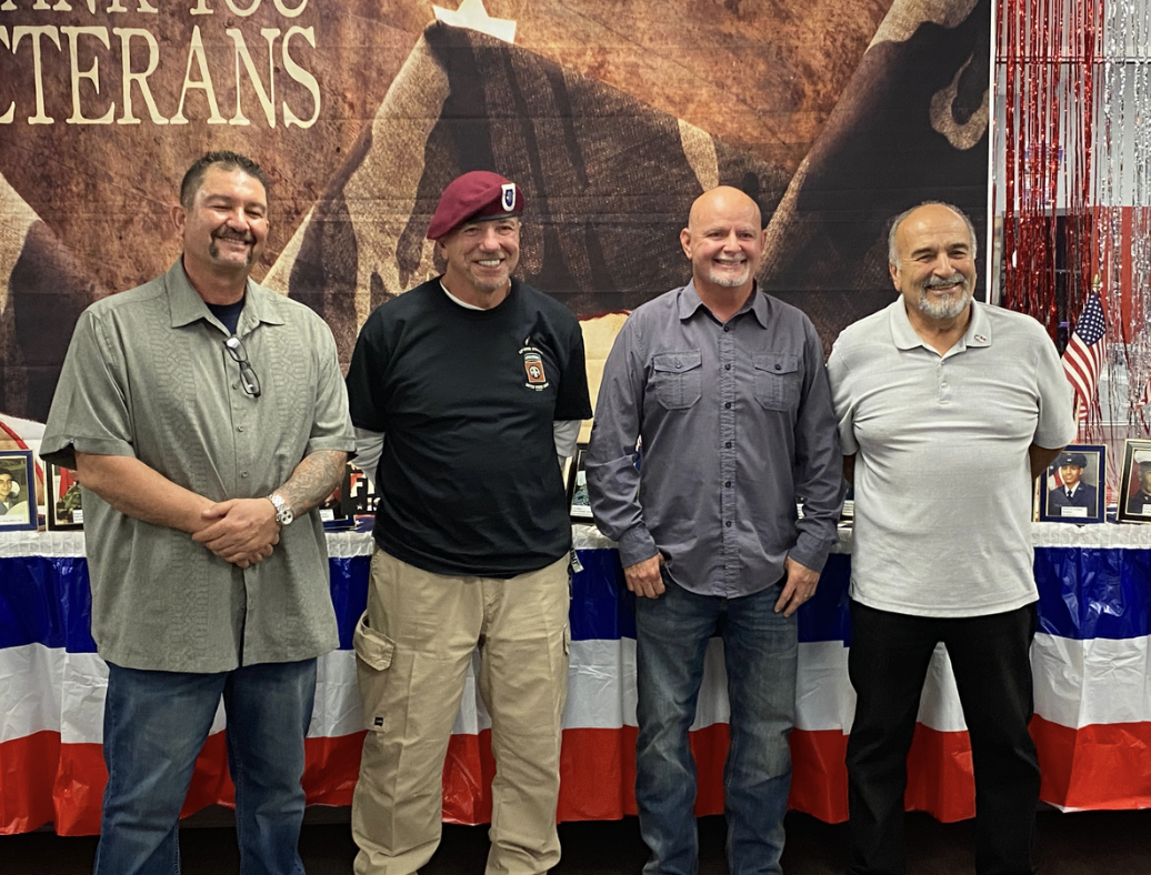 Four men standing in front of a patriotic display with a table decorated in red, white, and blue, featuring framed photographs and small American flags, at a Veterans event.