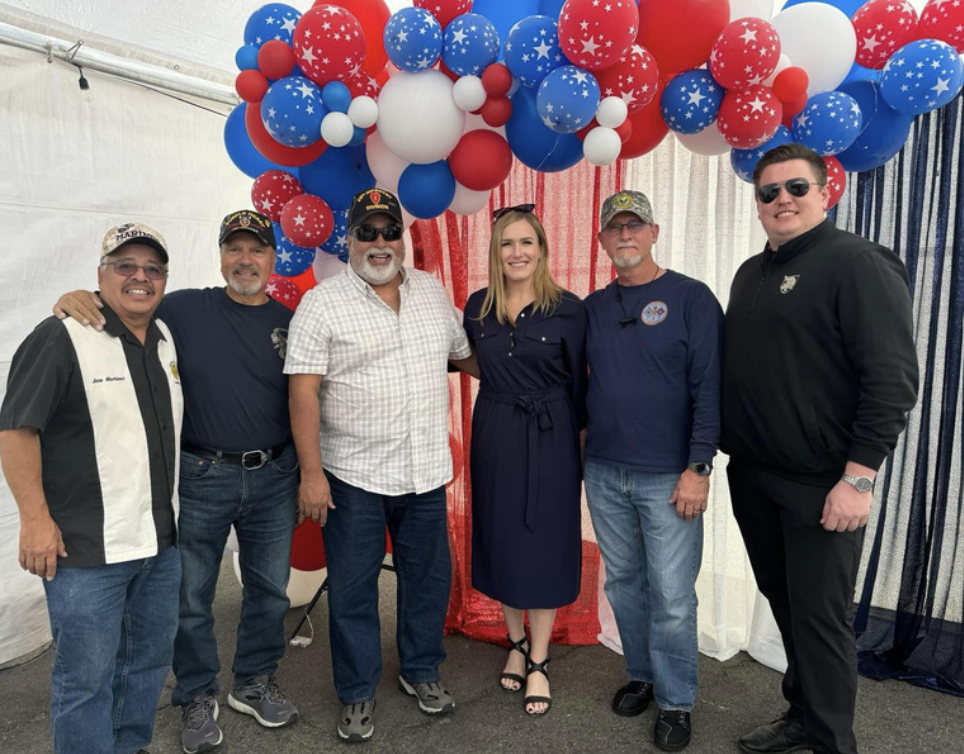 Six individuals posing in front of a patriotic balloon arch with red, white, and blue balloons and star patterns, inside a tent