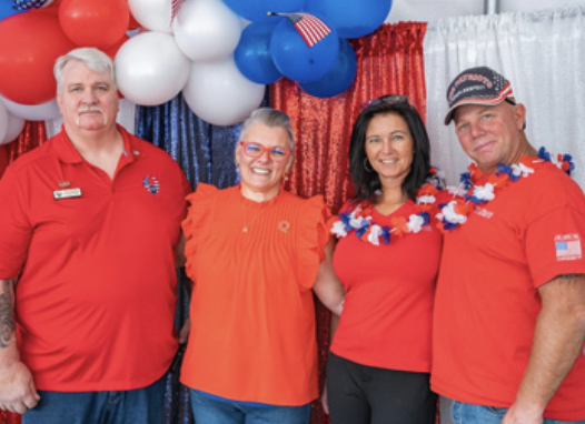 Four people standing in front of patriotic decorations, wearing red clothing and accessories, smiling for the camera.