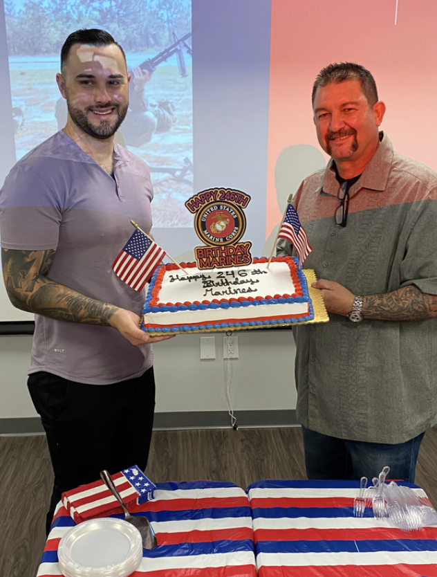 Two men holding a birthday cake decorated with American flags, with a Marine Corps badge and a message for a Marine's birthday. The birthday person is on the left, smiling, and the man on the right is also smiling. A table with a red, white, and blue