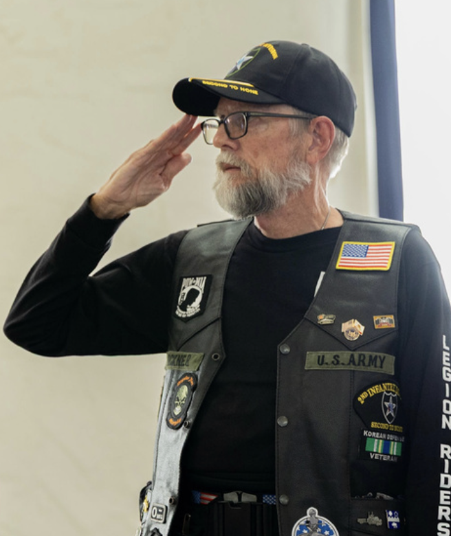 An older man with a gray beard and glasses salutes, wearing a black U.S. Army vest with various badges and patches, including an American flag, and a black cap with a military emblem.