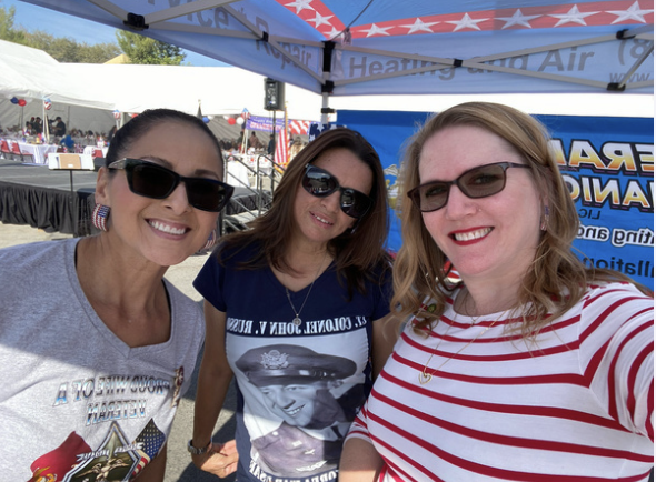 Three women smiling and taking a selfie at an outdoor event under a blue tent, with flags and a crowd in the background.