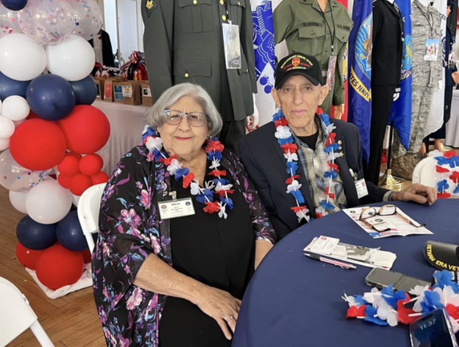 An elderly woman and man sitting at a table decorated with red, white, and blue flowers and leis, likely at a patriotic event or celebration. The woman wears glasses and a floral jacket, while the man wears a military cap and a dark blazer. Behind th