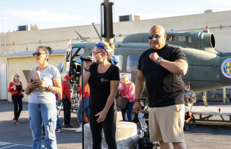 People gathered outdoors near a military helicopter, with one woman speaking into a microphone and others standing nearby, wearing casual clothing.