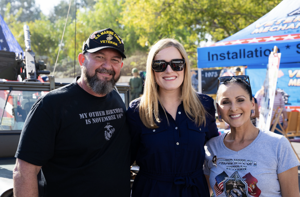 Three smiling people at an outdoor event, one man wearing a black cap and T-shirt with military theme, two women wearing sunglasses and T-shirts supporting veterans, standing in front of a blue tent with installation and sign, with trees and a vehicl