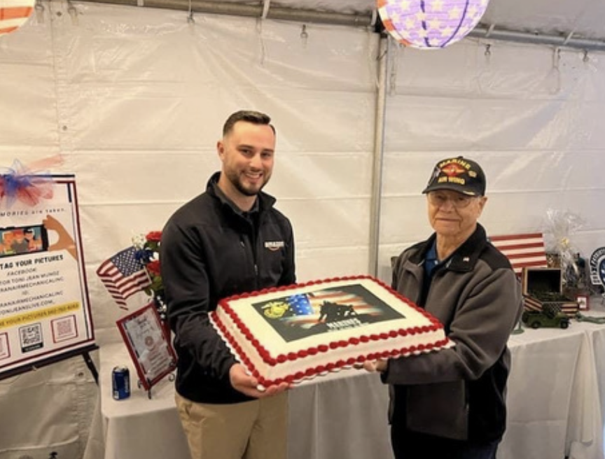 Two men holding a decorated cake with a military and patriotic theme, including an American flag and a military emblem, in a tent decorated for a celebration or event.