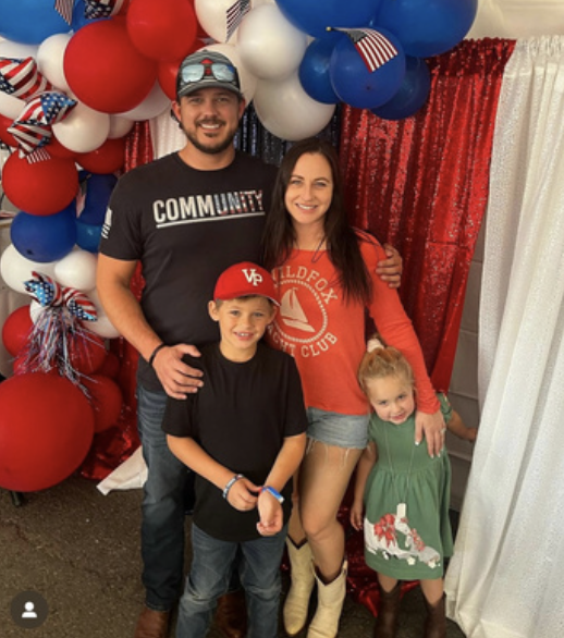A family of four standing in front of a patriotic red, white, and blue balloon backdrop with a red curtain and white curtain. They are smiling, with the man wearing a black t-shirt that says 'Community,' the woman wearing a red shirt, the boy in a bl