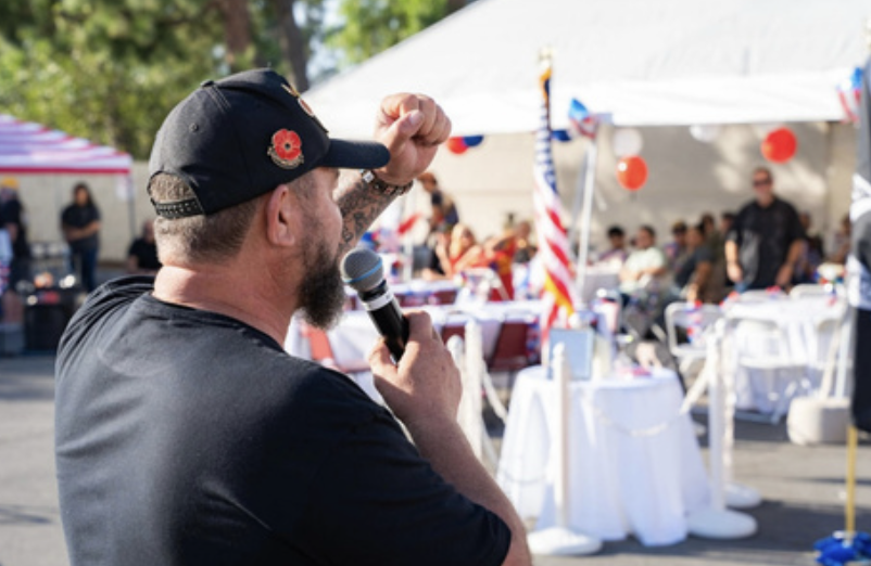 A man speaking into a microphone at an outdoor event with American flags and red, white, and blue decorations.