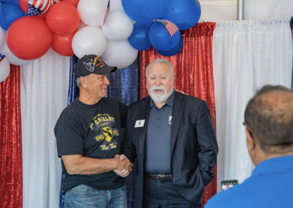 Two men shaking hands at a patriotic-themed event with red, white, and blue balloons and curtains in the background.