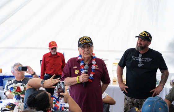 A woman in a maroon shirt and a navy cap with yellow lettering speaking into a microphone at a gathering, with people around her taking photos and listening, under a white tent.