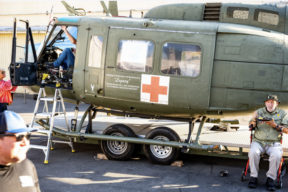 A military helicopter dressed as a medical aid vehicle, with a red cross symbol and sign referencing Vietnam veterans' legacy. People are gathered around, one person in a wheelchair holding a tray and another on the pilot seat.