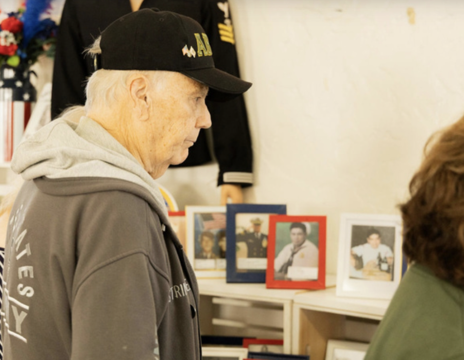 An elderly man wearing a black cap and gray hoodie looking at framed photographs on a shelf.