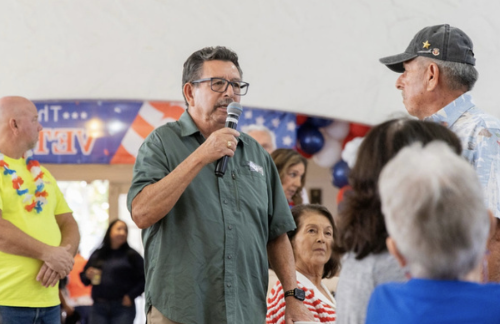 Man in green shirt holding a microphone speaking to a group of people at a patriotic event decorated with American flags and red, white, and blue balloons.